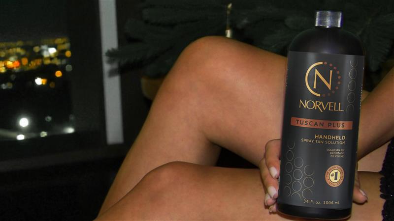 Person holding a bottle of Norvell Tuscan Plus handheld spray tan solution with a blurred background.