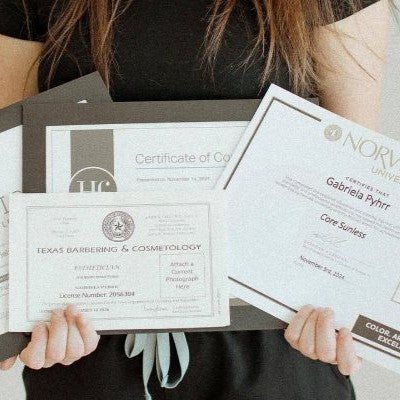 A person holds up several certificates and documents, including a Texas Barbering & Cosmetology certificate and a Norvell University certificate
