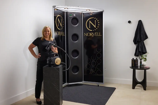 Spray tan artist with equipment in hand standing next to a large spray tan booth with 'Norvell' branding.