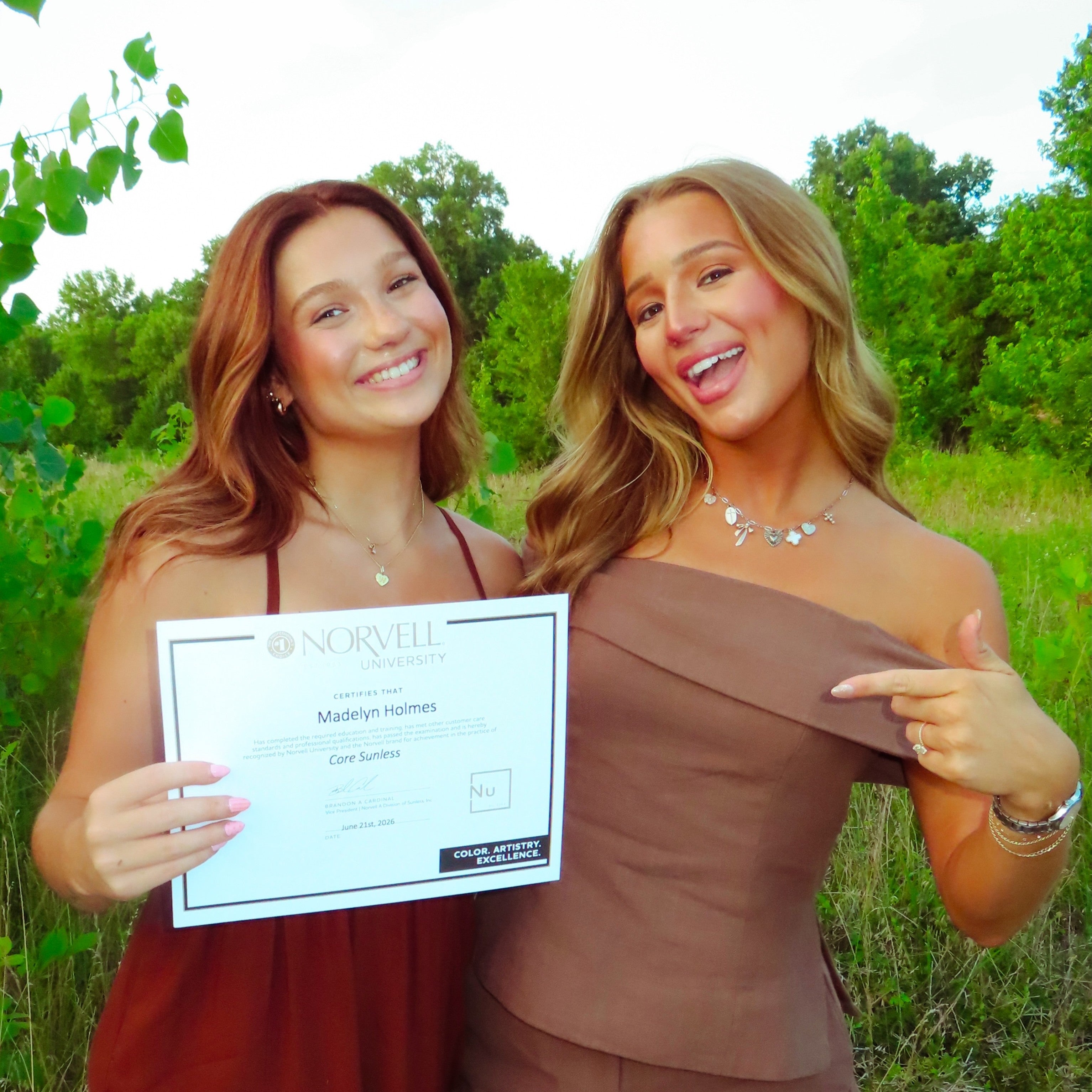 Two spray tan artists celebrating receiving their Norvell Spray Tan Artist certification in a lush green field. 