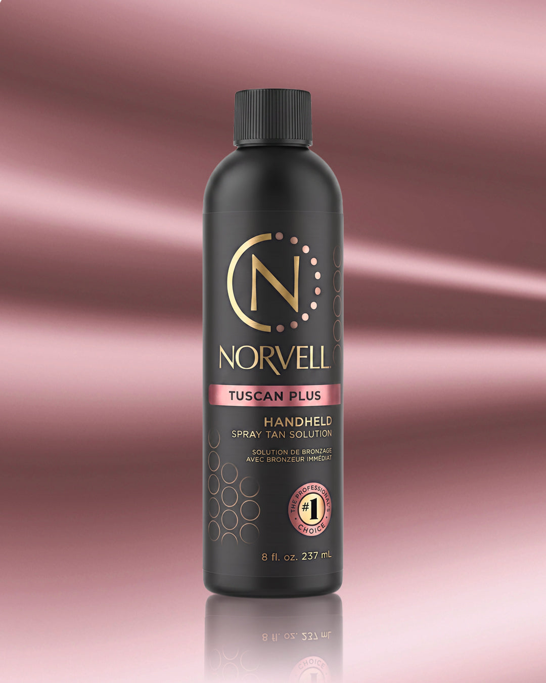 A bottle of Norvell Tuscan Plus Handheld Spray Tan Solution, featuring a matte black bottle with gold and pink accents, set against a pink gradient background with a soft reflection.